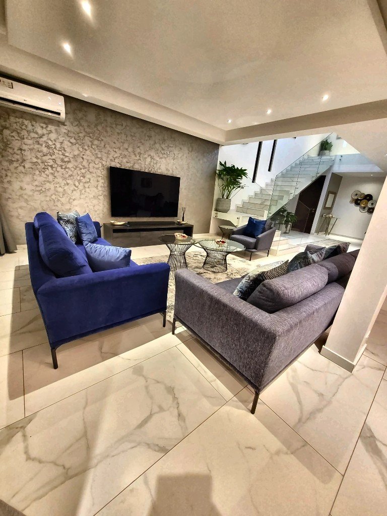 Luxury living room with blue velvet and grey seating, glass coffee tables, marble floors, and glass-railed staircase — D'anges Interiors
